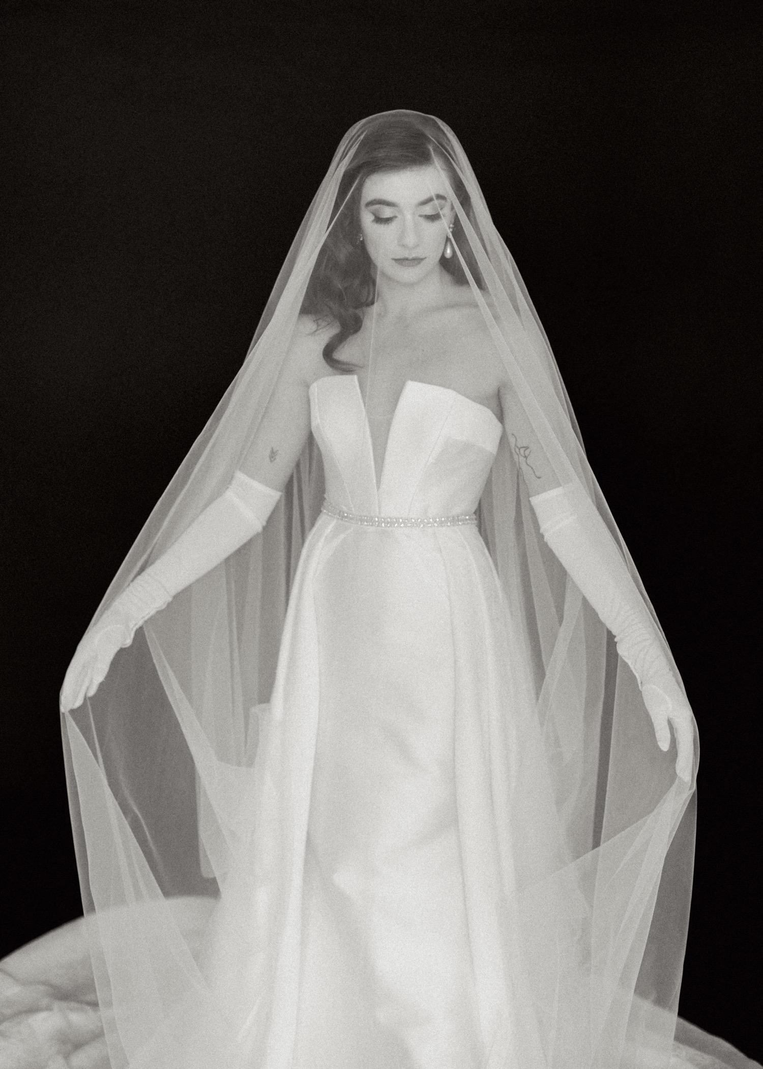 https://fetch.getnarrativeapp.com/static/cbb3543c-76b2-4397-ba8b-8abac010d767/Bride-in-white-sleeveless-wedding-dress,-overskirt,-gloves,-and-long-veil-during-studio-bridal-session-with-black-backdrop.jpg?w=1500