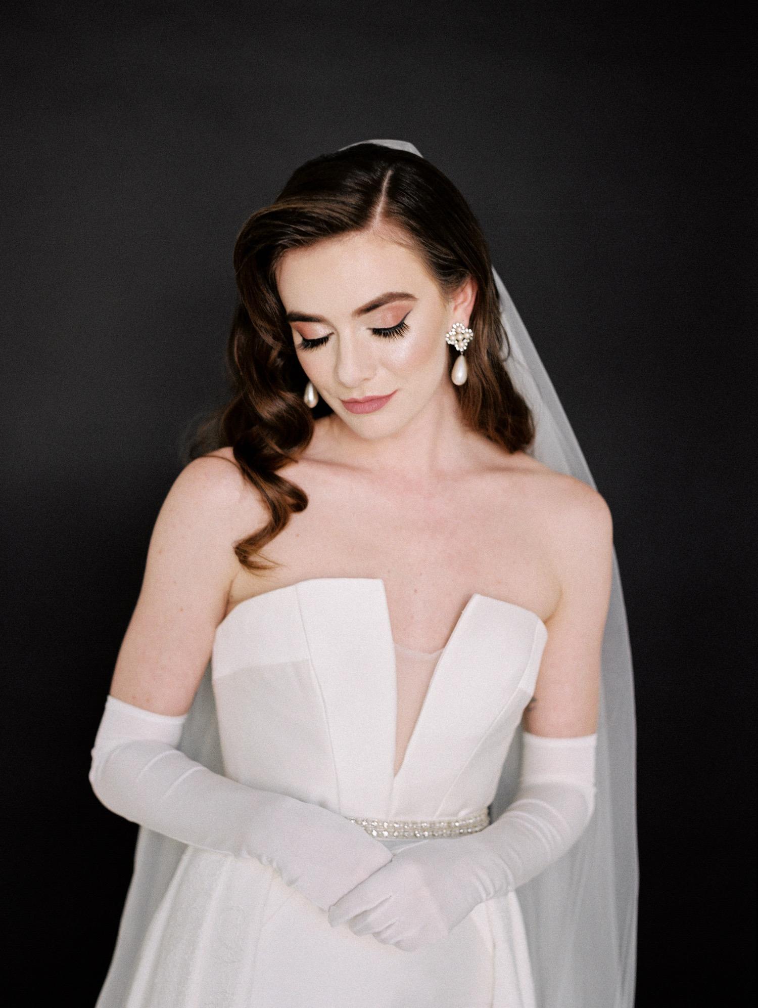 https://fetch.getnarrativeapp.com/static/e523c136-3c84-4c74-a4a2-b711ce1c8273/Smirking-bride-in-white-wedding-dress,-overskirt,-gloves,-and-veil-during-studio-bridal-session-with-black-backdrop.jpg?w=1500